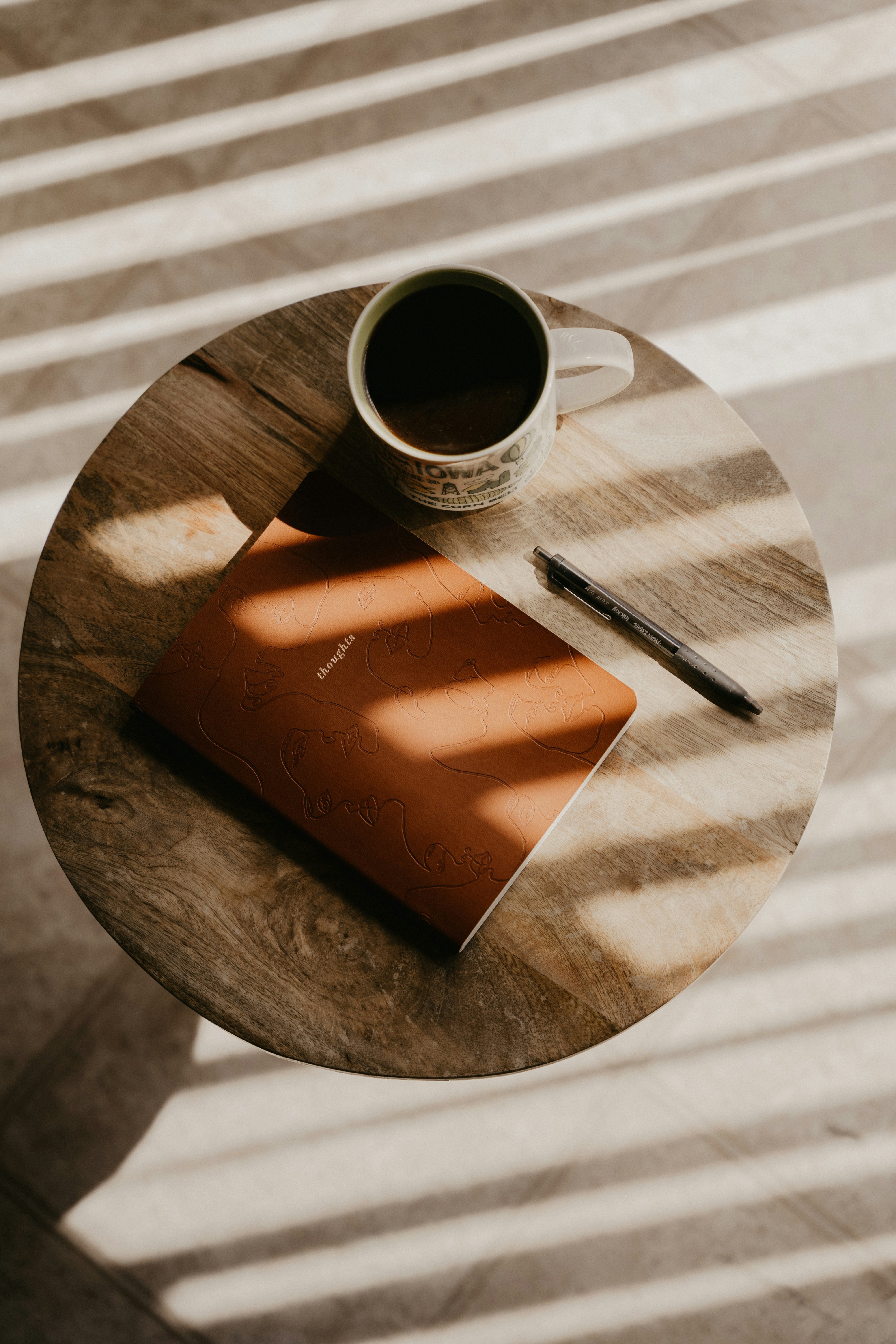 mesa con un cuaderno, boligrafo y una taza de cafe, la luz entra tenue por la ventana generando sombras sobre la mesa